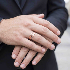 4 of 8: Close view of a person in a dark suit wearing a polished chased copper bypass ring on the ring finger. The open ends overlap across the top, showing the hand applied linear texture and warm rose copper color.