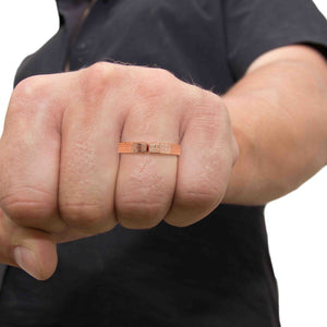 6 of 8: Close-up of a man’s hand wearing a 6mm linen textured copper cuff ring on the ring finger. The open band design features a small gap at the top and a woven fabric pattern engraved across the outer surface. The solid copper ring has a warm rose gold tone with a polished finish. The hand is shown in a relaxed fist position against a clean white background, highlighting the ring’s unisex style and everyday wear appeal.
