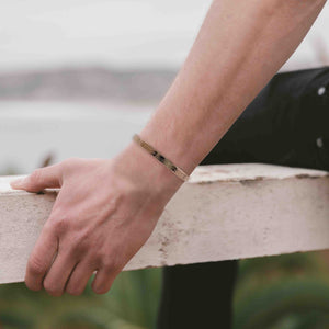 6 of 9: A close-up image of a man wearing a 5mm wide bronze cuff bracelet with a bark texture on his wrist. The bracelet has a natural golden-brown tone and an open-ended design. He is leaning casually on a white concrete surface outdoors, with a blurred landscape and water in the background, giving the photo a relaxed, coastal vibe.