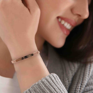 4 of 9: A close-up image of a smiling woman wearing a 5mm wide bronze cuff bracelet with a hand-applied bark texture. The bracelet rests on her wrist, showing off its warm bronze tone and subtle oxidized detailing. She is dressed in a cozy gray knit sweater and white top, creating a soft, casual look that highlights the bracelet’s natural charm.