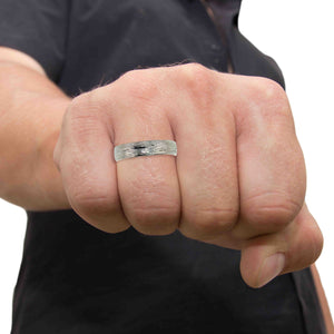 2 of 9: A close-up image of a person's clenched fist wearing a 5mm wide stainless steel ring with a bark-textured finish. The ring has a polished, domed surface on the inside and features a rugged, hand-carved bark pattern on the outside. The ring is worn on the middle finger, and the wearer is dressed in a dark short-sleeved shirt. The background is removed, isolating the subject against a plain white backdrop, which highlights the ring’s texture and reflective surface.