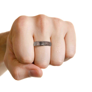 8 of 9: A close-up image of a clenched fist wearing a 5mm wide stainless steel ring with a bark-textured finish. The ring is positioned on the middle finger and features a hand-carved, organic pattern on the outer surface with a smooth, domed, polished interior for a comfort fit. The hand is photographed against a plain white background, emphasizing the contrast between the ring’s rugged texture and its shiny finish. The image highlights the ring’s unisex style and natural, handcrafted design.