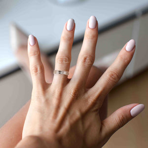 4 of 9: A close-up image of a woman's hand wearing a 5mm wide stainless steel ring with a bark-textured finish on her ring finger. The ring has a polished, domed interior for a comfort fit and a hand-carved bark pattern on the outer surface. Her nails are neatly manicured and painted in a soft, pale pink color, with one accent nail featuring a glittery finish. The background is softly blurred, showing part of her legs and high-heeled shoe, suggesting a stylish indoor setting. The lighting highlights the ring's text