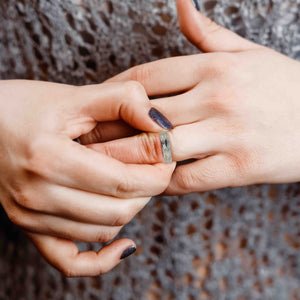 6 of 9: A close-up image of a person’s hands as they gently slide a stainless steel ring onto their finger. The ring is 5mm wide with a hand-carved bark texture on the outside and a polished, domed interior for a comfort fit. The individual has dark, glittery purple nail polish and is wearing a gray lace-patterned top, adding a touch of elegance to the scene. The lighting softly highlights the textured surface of the ring and the natural tones of the skin, creating a warm and intimate atmosphere.