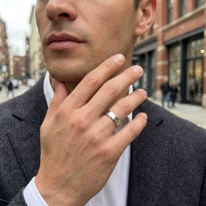 4 of 8: Man dressed in a dark coat wearing a polished 5mm chased stainless steel comfort fit ring on his ring finger. His hand is raised near his chin on a city street, showing the reflective silver tone band in an everyday setting.