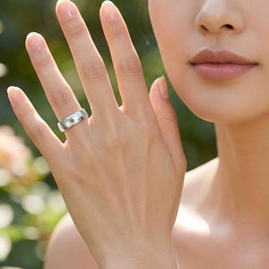 2 of 8: Close view of a woman holding her hand near her face while wearing a polished 5mm chased stainless steel comfort fit ring on her ring finger. The reflective silver tone band stands out against a softly blurred green outdoor background.