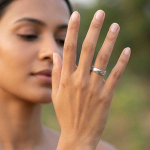 6 of 8: Woman holding her hand in front of her face while wearing a polished 5mm chased stainless steel comfort fit ring on her ring finger. The reflective band is shown outdoors against a softly blurred green background.
