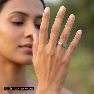4 of 8: Woman holding her hand in front of her face while wearing a polished 5mm chased stainless steel comfort fit ring on her ring finger. The reflective band is shown outdoors against a softly blurred green background.  AI Generated Model.
