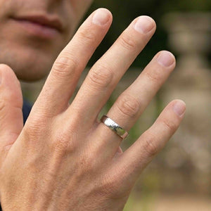 7 of 8: Close view of a man wearing a polished 5mm chased stainless steel comfort fit ring on his ring finger. The silver tone band catches the light while his hand is raised near his face against a softly blurred outdoor background.