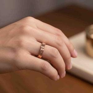 2 of 8: Close-up of a hand wearing a 5mm linen textured copper cuff ring on the ring finger. The open band design shows a small gap at the top, highlighting the hand applied woven pattern on the outer surface. The copper has a warm rose gold tone with a polished finish. The hand rests naturally on a wooden surface with a softly blurred background, emphasizing the ring’s texture and everyday wear style.