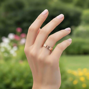 8 of 9: Close-up outdoor lifestyle image of a hand wearing a 6mm solid bronze cuff ring with a bark-textured finish. The open band design features a small gap at the top, highlighting its adjustable style. The warm coppery bronze contrasts softly with the wearer’s light skin tone and pale pink manicured nails. A blurred garden background with green foliage and soft flowers creates a fresh, natural setting.