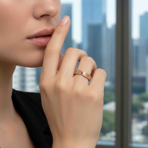 6 of 9: Close-up lifestyle image of a woman wearing a 6mm solid bronze cuff ring with a bark-textured finish. The open band design shows a small gap at the top, highlighting its adjustable style. The warm coppery bronze contrasts softly with her light skin tone. She gently touches her lips with her index finger, and a blurred city skyline is visible in the background, creating a modern, sophisticated setting.