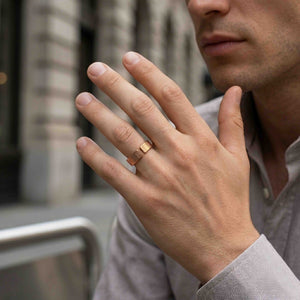 6 of 9: Man wearing a 6mm chased bronze cuff ring on his finger while holding his hand near his face in an urban setting. The polished bronze ring has an open cuff shape and a hand applied chased texture with narrow vertical marks.