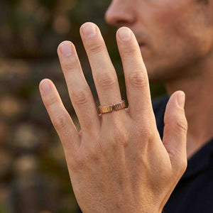 4 of 9: Man wearing a 6mm chased bronze cuff ring outdoors with his hand raised in front of a softly blurred background. The open ends of the polished bronze band face forward, showing the hand applied vertical texture and unisex fit.