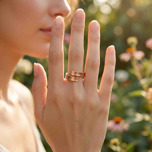 8 of 9: Hand wearing the 5.5mm bark copper bypass ring in a garden setting with blurred flowers, showing the warm polished copper, hand applied bark texture, open wrap style, and easy everyday look.