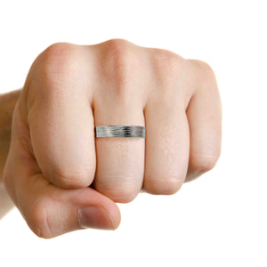 4 of 8: Close up of a hand wearing a 7mm stainless steel flat band ring with a hand carved bark texture. The ring is shown on a finger of a gently clenched fist, highlighting its flat edges and polished finish. The organic bark pattern runs across the center of the band, reflecting light with subtle grooves and texture. The interior appears smooth and shiny. The background is clean white, emphasizing the modern unisex design.