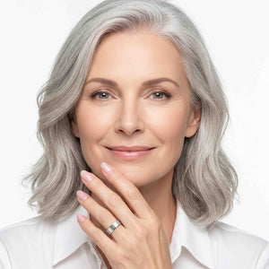 8 of 9: Polished stainless steel flat band ring worn on a woman’s finger as she rests her hand near her chin, featuring a wide smooth profile, reflective silver finish, and clean modern style with a white shirt and soft white background.
