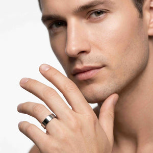 6 of 9: Polished stainless steel flat band ring worn on a man’s finger near his face, featuring a wide smooth profile, reflective silver finish, flat surface, and modern unisex style against a clean white background.