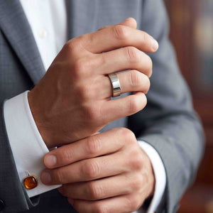 2 of 9: Polished stainless steel flat band ring worn on a man’s finger as he adjusts a white shirt cuff under a gray suit jacket with amber cufflink, creating a refined formal look.