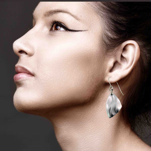 6 of 7: Side profile close up of a woman wearing a brushed aluminum flame petal dangle earring. The lightweight silver tone earring features curved die struck detailing and hangs from a stainless steel French ear wire. The sculpted petal shaped drop reflects light with a soft satin finish. The model has dark hair pulled back and wears dramatic winged eyeliner against a dark neutral background.