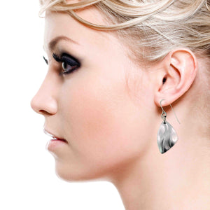 2 of 7: Side profile of a woman with blonde hair wearing a brushed aluminum flame petal dangle earring. The lightweight silver tone earring features curved die struck detailing and hangs from a stainless steel French ear wire. The sculpted petal shape drops just below the earlobe and reflects light with a soft satin finish against a clean white background.