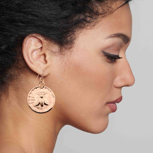 6 of 7: Close-up of a woman wearing Bronze Queen Bee Disc Drop Earrings with a detailed design.