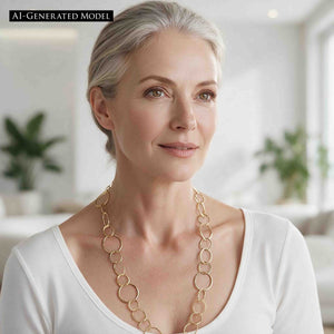 4 of 8: AI Generated Model with gray hair wearing the Chased Nu Gold Brass Chain Link Necklace over a white scoop neck top. The long golden necklace frames the neckline with mixed round and oval links in several sizes, showing how the hand hammered brass design looks with a clean, classic outfit.