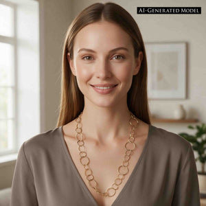 2 of 8: AI Generated Model wearing the Chased Nu Gold Brass Chain Link Necklace with a taupe V neck top in a softly lit interior. The 26 inch necklace falls along the neckline and features mixed round and oval Nu Gold brass links in three sizes, creating an open, airy look with warm golden color.