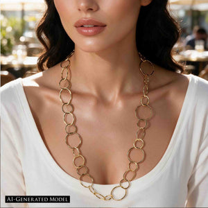 6 of 8: AI Generated Model wearing the Chased Nu Gold Brass Chain Link Necklace with a white scoop neck top at an outdoor cafe. The long golden necklace features mixed round and oval links in several sizes, with hand hammered texture that catches the light against the simple white neckline.