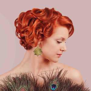 4 of 7: Woman with styled red hair wearing a peacock feather garment against a pink background wearing Christmas Starburst Nu Gold Brass Earrings