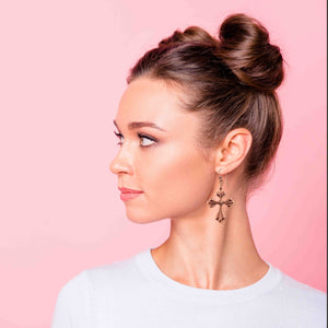 4 of 7: Woman with styled hair and wear Copper Baroque Cross Drop Earrings against a pink background