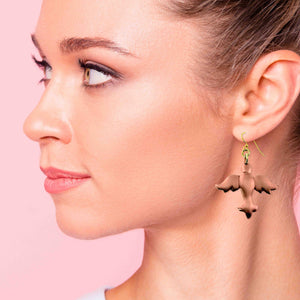 2 of 6: Close-up of a woman wearing Copper Christmas Dove Dangle Earrings against a pink background