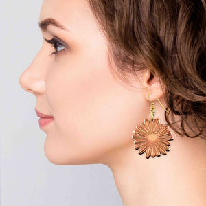 6 of 7: Side profile of a woman with short brown hair wearing a handcrafted copper daisy dangle earring. The earring features detailed petal texture and a polished finish, suspended from a gold tone brass French hook. It hangs just below the earlobe, catching the light with a warm glow and showing its natural movement when worn.
