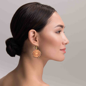 2 of 7: Side profile of a woman wearing handcrafted copper daisy dangle earrings with detailed petal texture and a polished finish. The earring hangs from a gold tone brass French hook, catching the light with a warm glow against her skin, showing its size and elegant movement when worn.