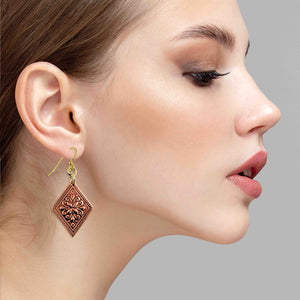 4 of 7: Side profile of a woman wearing a copper floral diamond dangle earring. The earring features a diamond shaped polished copper panel with raised ornate floral detailing and a decorative textured border. It hangs from gold tone brass French ear wires with small connecting rings. The warm raw copper finish softly reflects light as the earring dangles about 2 inches below the ear, highlighting its handcrafted design against the model’s pulled back hair and neutral background.