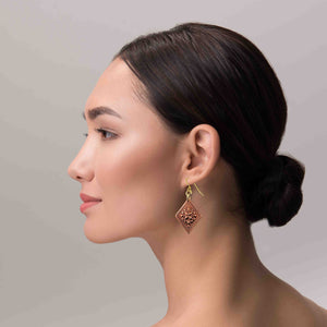6 of 7: Side profile of a woman wearing a copper floral diamond dangle earring. The earring features a polished diamond shaped copper panel with raised ornate floral detailing and a decorative textured border. It hangs from gold tone brass French ear wires with small connecting rings. The warm raw copper finish catches the light as the earring dangles below the ear, highlighting its handcrafted design against the model’s dark pulled back hair and neutral background.