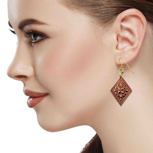 2 of 7: Side profile of a woman wearing a copper floral diamond dangle earring. The earring features an ornate raised floral pattern on a diamond shaped copper panel with a textured border. It hangs from gold tone brass French ear wires with small connecting rings. The polished copper design measures about 2 inches long and gently dangles below the ear, adding warm metallic shine against the model’s smooth skin and pulled back hair.