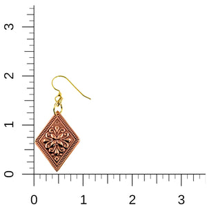 5 of 7: Copper floral diamond dangle earring displayed next to a measurement ruler for scale. The earring features a diamond shaped polished copper panel with raised ornate floral detailing and a textured border. It hangs from gold tone brass French ear wires with connecting rings. The ruler shows the earring measures approximately 2 inches long and about 7/8 inch wide, highlighting its lightweight handcrafted design.