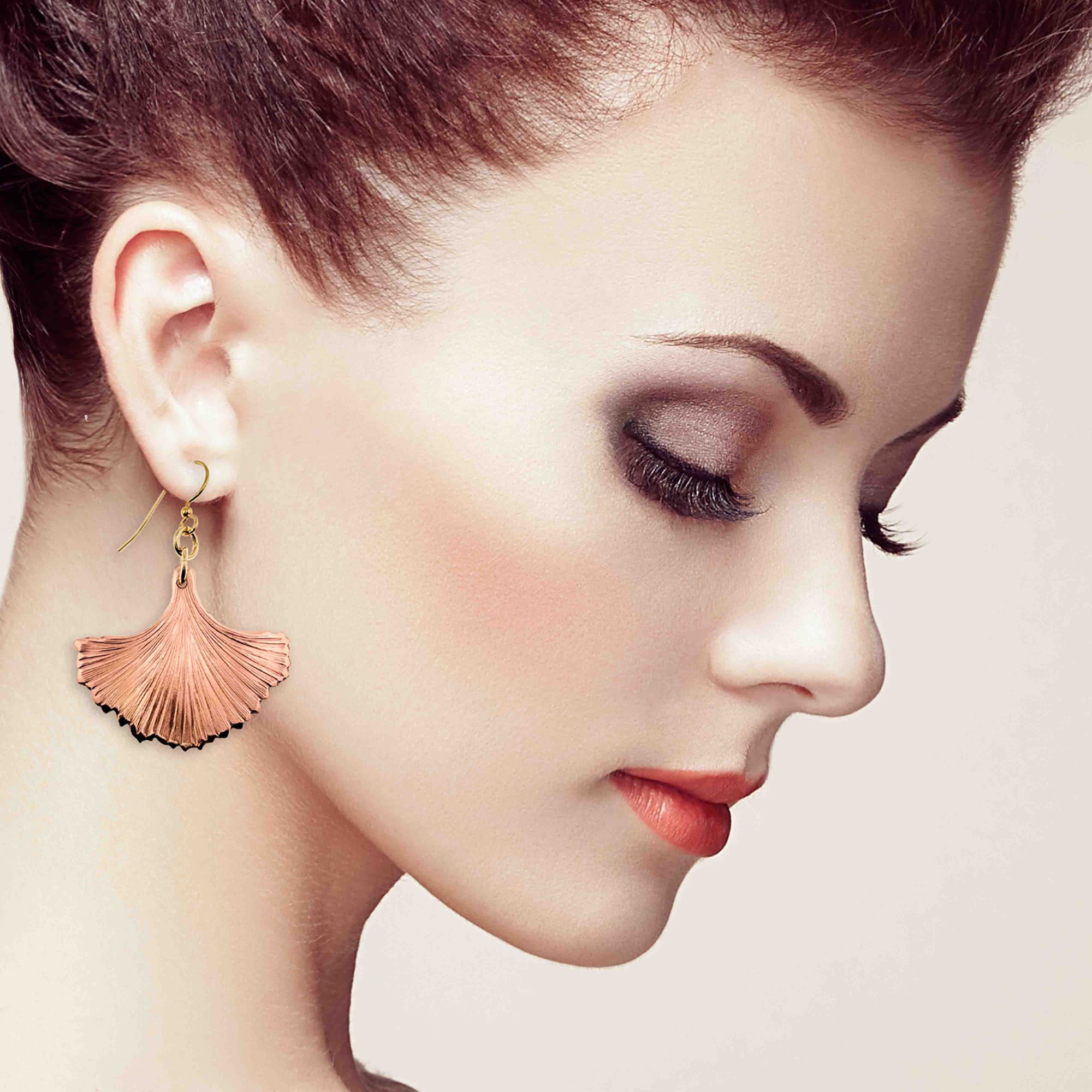 Side profile of a woman with fair skin and swept-back auburn hair, wearing dramatic eye makeup and coral lipstick, shown with her eyes closed while modeling a large copper ginkgo leaf dangle earring with fine radiating grooves and darkened tips, hanging from a gold tone French hook against a soft beige background.