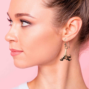 2 of 7: A woman with her hair tied back is wearing a handcrafted copper lily flower drop earring. The earring features a detailed, flowing petal design with a warm copper finish, hanging from a copper French ear wire. The background is soft pink, highlighting the natural tones of her skin and the earring’s metallic shine.