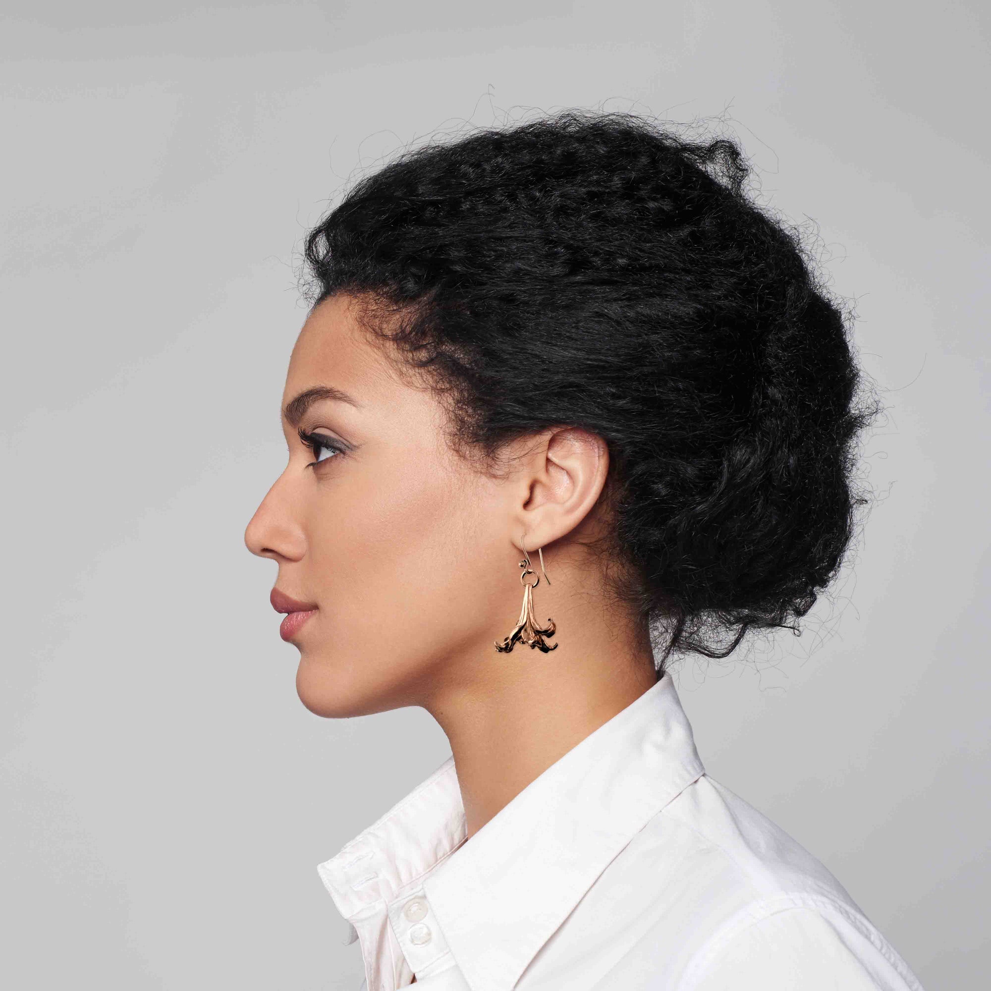 Side profile of a woman with dark curly hair in a low bun, wearing a white collared shirt and long copper lily flower drop earrings, shown against a light gray background.