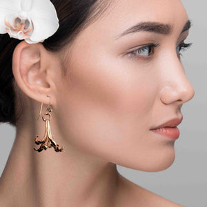 6 of 7: A woman with smooth, radiant skin and dark hair styled back is wearing a copper lily flower drop earring. The earring features a detailed petal design and hangs from a copper French ear wire. A white orchid flower is tucked behind her ear, adding a soft, tropical touch to the elegant, natural look. The background is a neutral gray, drawing focus to the earring and her serene expression.