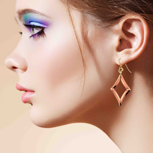 6 of 7: Close up side profile of a woman wearing a handcrafted copper open diamond drop earring. The polished geometric copper frame hangs from a brass French ear wire, catching the light against her skin. She has soft pink lips and bold eye makeup in shades of blue and purple, with her hair pulled back, highlighting the warm metallic tone and modern minimalist design of the earring.