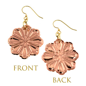 3 of 7: Front and back view of handcrafted copper flower dangle earrings shown against a light neutral background. The front displays polished, die struck petal detailing with raised texture and a warm rosy copper shine. The back shows a smooth polished copper surface. Each earring hangs from gold tone jump rings and brass French hook ear wires. Floral design measures about 2 inches long.