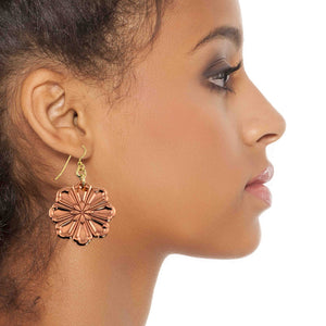 2 of 7: Close up side profile of a woman wearing a handcrafted copper flower dangle earring. The polished copper bloom features raised, die struck petal detailing and hangs about 2 inches below the earlobe from a brass French hook ear wire. The warm rosy copper tone contrasts softly with her skin, showing the scale and elegant movement of the floral design.
