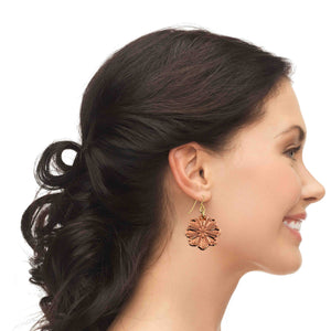 6 of 7: Side profile of a smiling woman wearing a polished copper flower dangle earring. Her dark hair is styled in a low, softly curled updo. The handcrafted earring features a eight petal bloom with raised, die struck detailing and a warm rosy copper shine. It hangs about 2 inches below the earlobe from a brass French hook ear wire, showing scale and graceful movement against a light background.