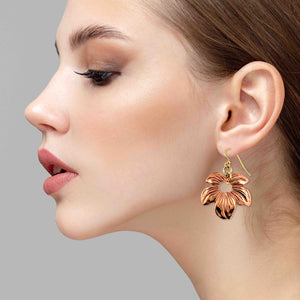 6 of 7: Side profile of a woman wearing a copper petal blossom dangle earring with polished floral detailing and an open center, suspended from a brass French ear wire, shown against a soft neutral background.