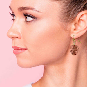 2 of 7: Close-up of a woman wearing a Copper Pine Cone Dangle Earring against a pink background