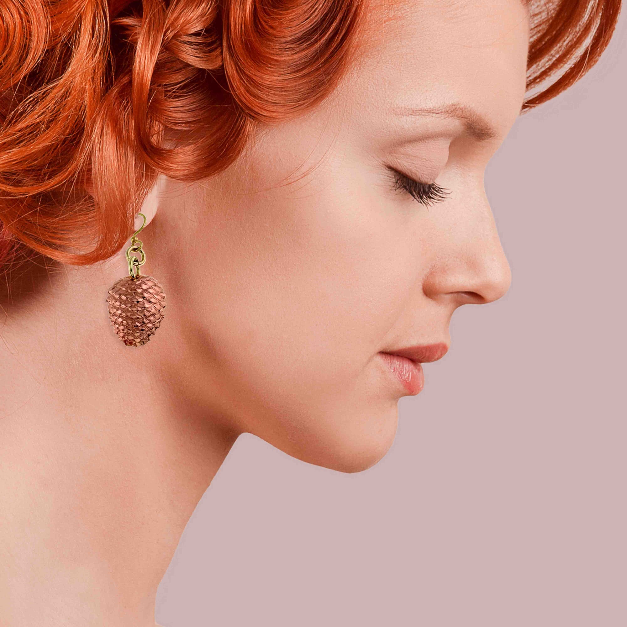 Profile of a woman with red hair wearing Copper Pine Cone Dangle Earrings on a pink background