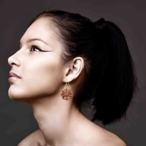 6 of 7: Woman shown in side profile wearing copper sunburst flower dangle earrings. The polished copper floral charm features layered petal detailing in a radiant sunburst design and hangs from a brass French ear wire. The warm copper tone contrasts with the model’s smooth skin and dark hair, highlighting the earring’s elegant drop length and decorative texture against a soft dark background.
